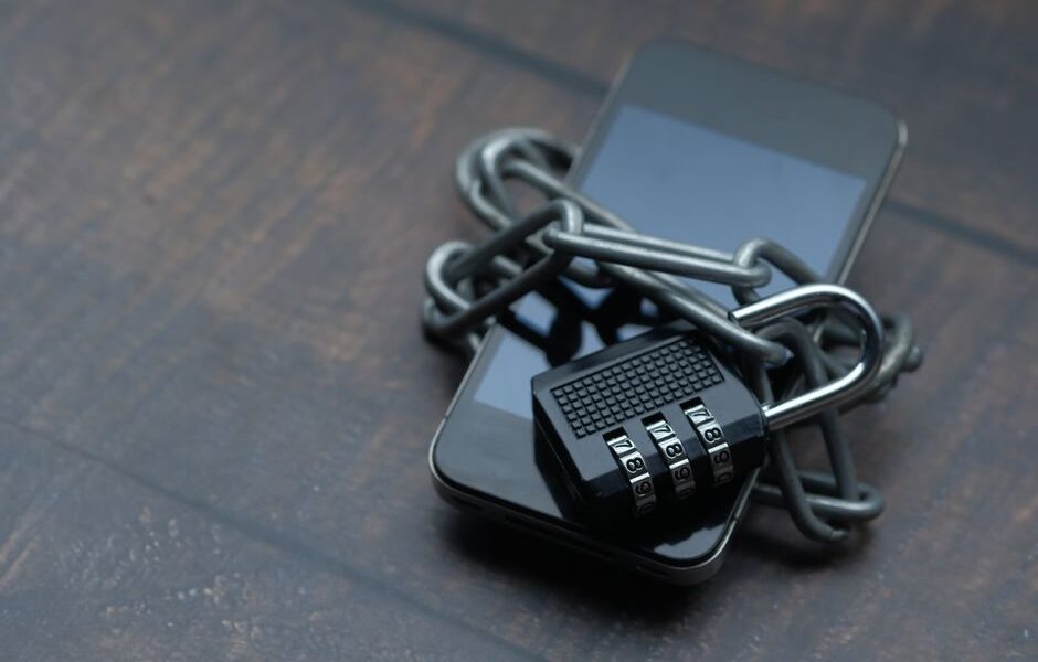 Smartfon owinięty metalowym łańcuchem z zamkniętą kłódką szyfrową na drewnianym blacie.