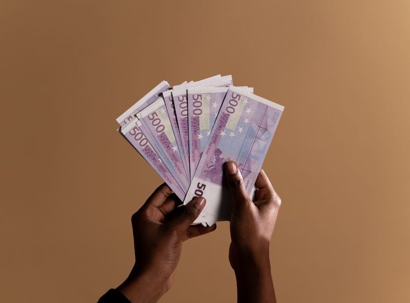 Dłonie trzymające wachlarz banknotów o nominale pięciuset euro na brązowym tle.