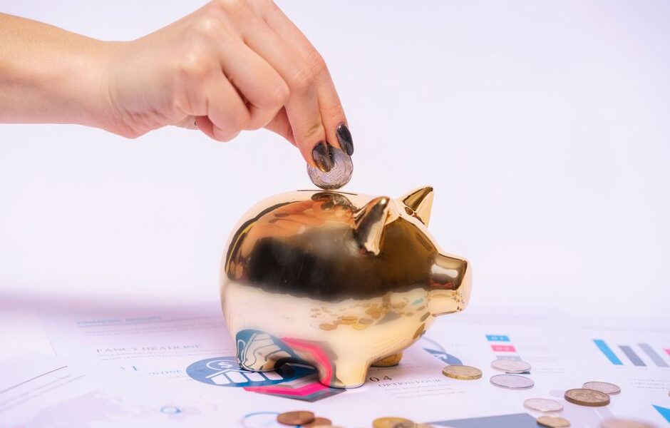 Dłoń wrzuca monetę do złotej skarbonki stojącej na wykresach finansowych.