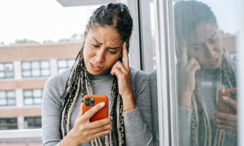Kobieta z warkoczykami trzymająca telefon i dotykająca dłonią skroni przy oknie.