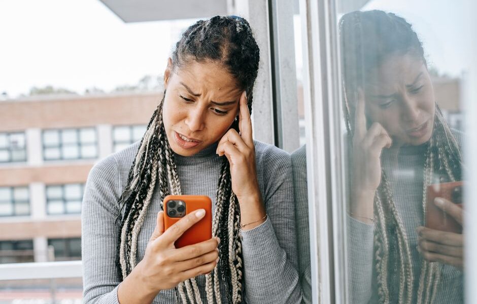 Kobieta z warkoczykami trzymająca telefon i dotykająca dłonią skroni przy oknie.