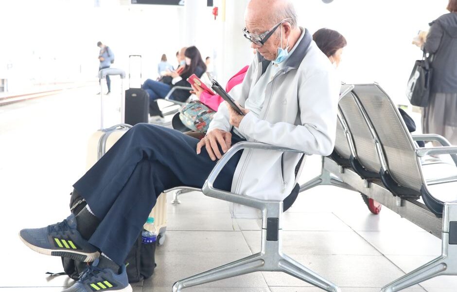 Starszy mężczyzna w okularach przegląda tablet siedząc na lotniskowej poczekalni.