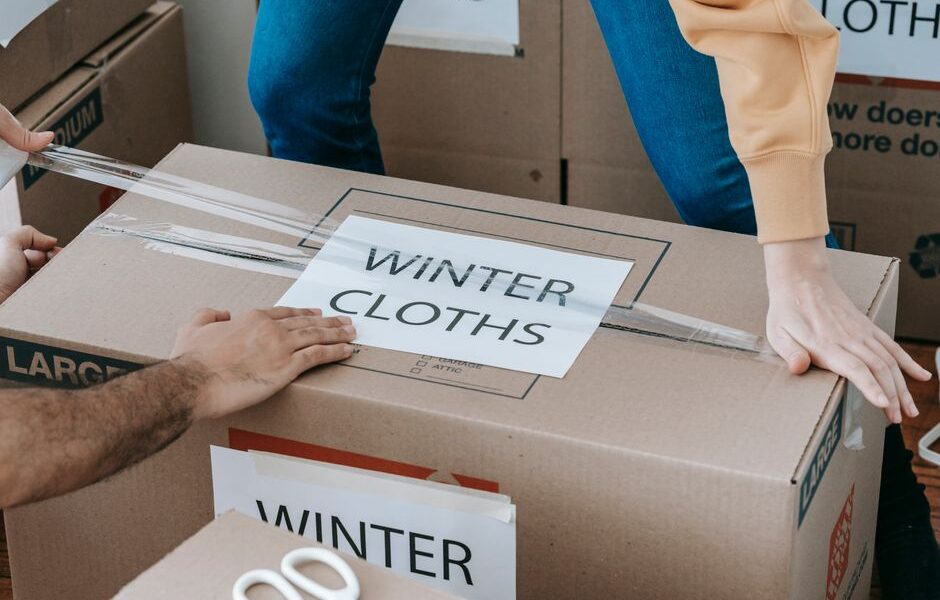 Osoby zaklejają taśmą kartonowe pudło z napisem Winter Cloths