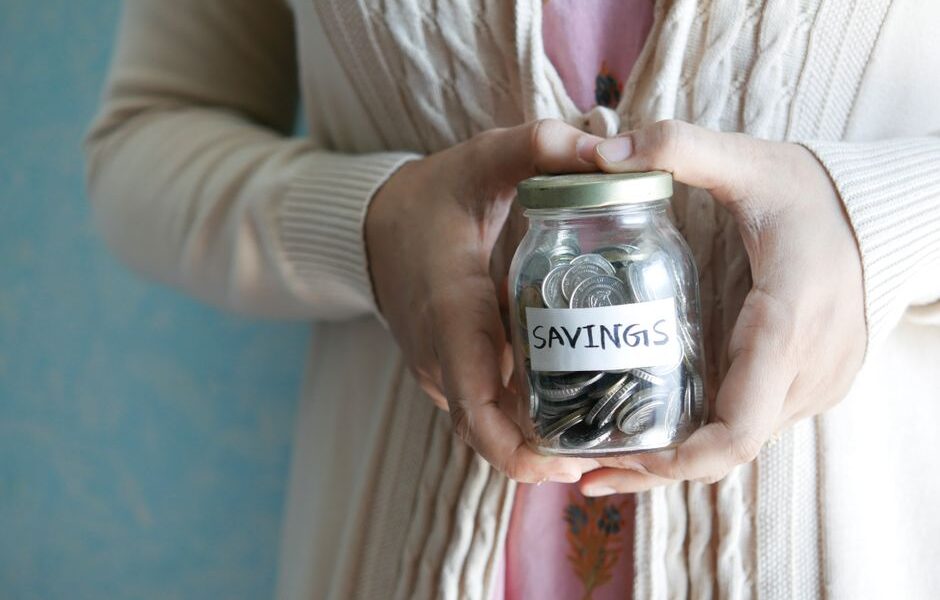 Dłonie trzymające szklany słoik z monetami i napisem SAVINGS.